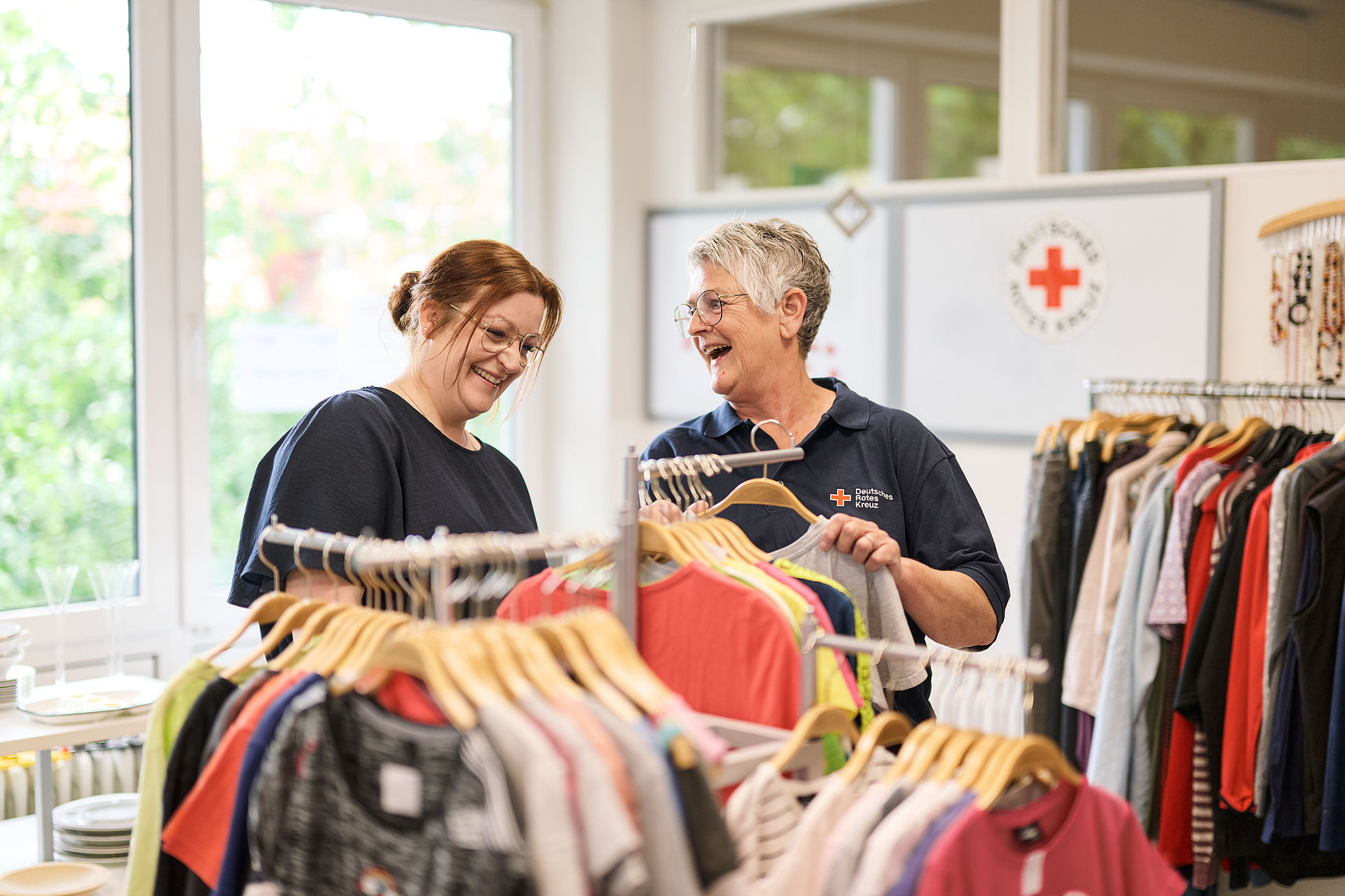 Zwei Frauen stehen an einem Kleiderständer und lachen, während Sie sich Kleidung anschauen
